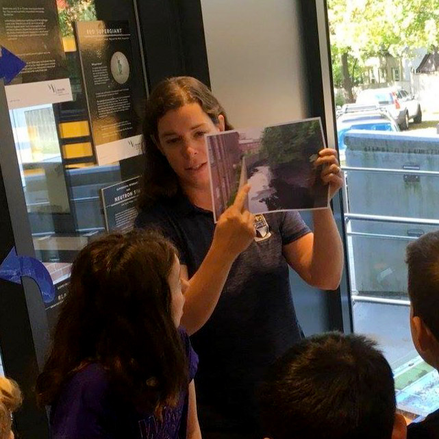 Kerry Reed A woman holds up and explains a photo of a waterway to a group of children during an educational session indoors, with natural light coming through large windows.