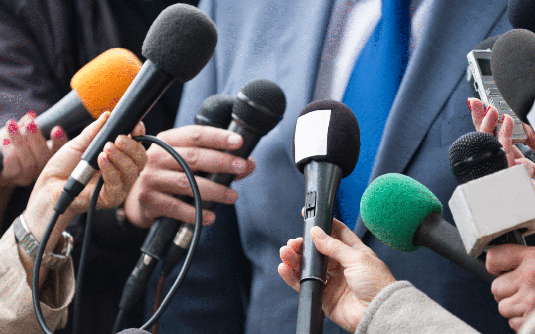 Close-up of multiple reporters holding out microphones toward a person in a suit and blue tie during a press interview.