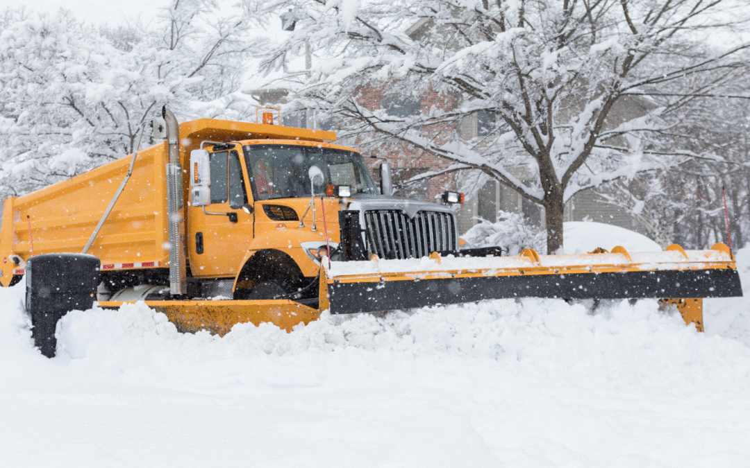 A large yellow municipal snowplow truck clears a residential street during heavy snowfall, pushing a wide path through deep snow. Snow-covered trees and houses line the background as steady snow continues to fall.